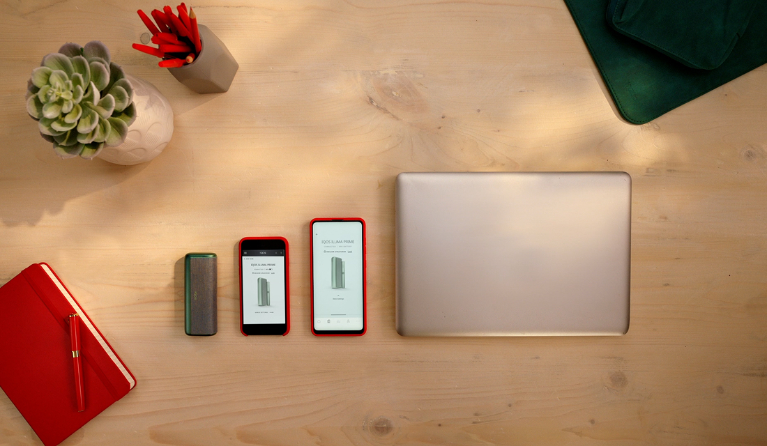 En IQOS ILUMA-enhet, två mobiltelefoner som visar IQOS-appen och en laptop på ett bord