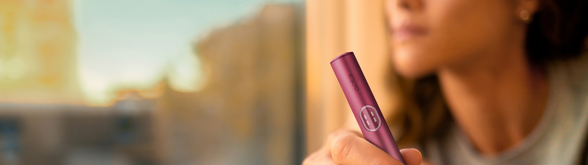 Kvinna som håller i en Garnet Red IQOS ILUMA i PRIME-enhet medan hon tittar ut genom fönstret