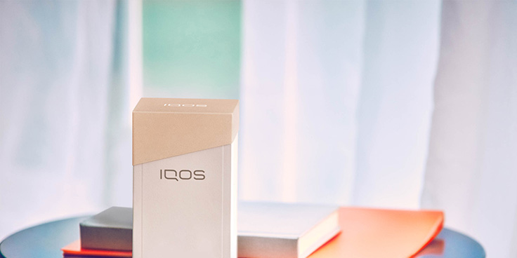 Blond woman checking her phone with a IQOS device on the table and a handbag next to her